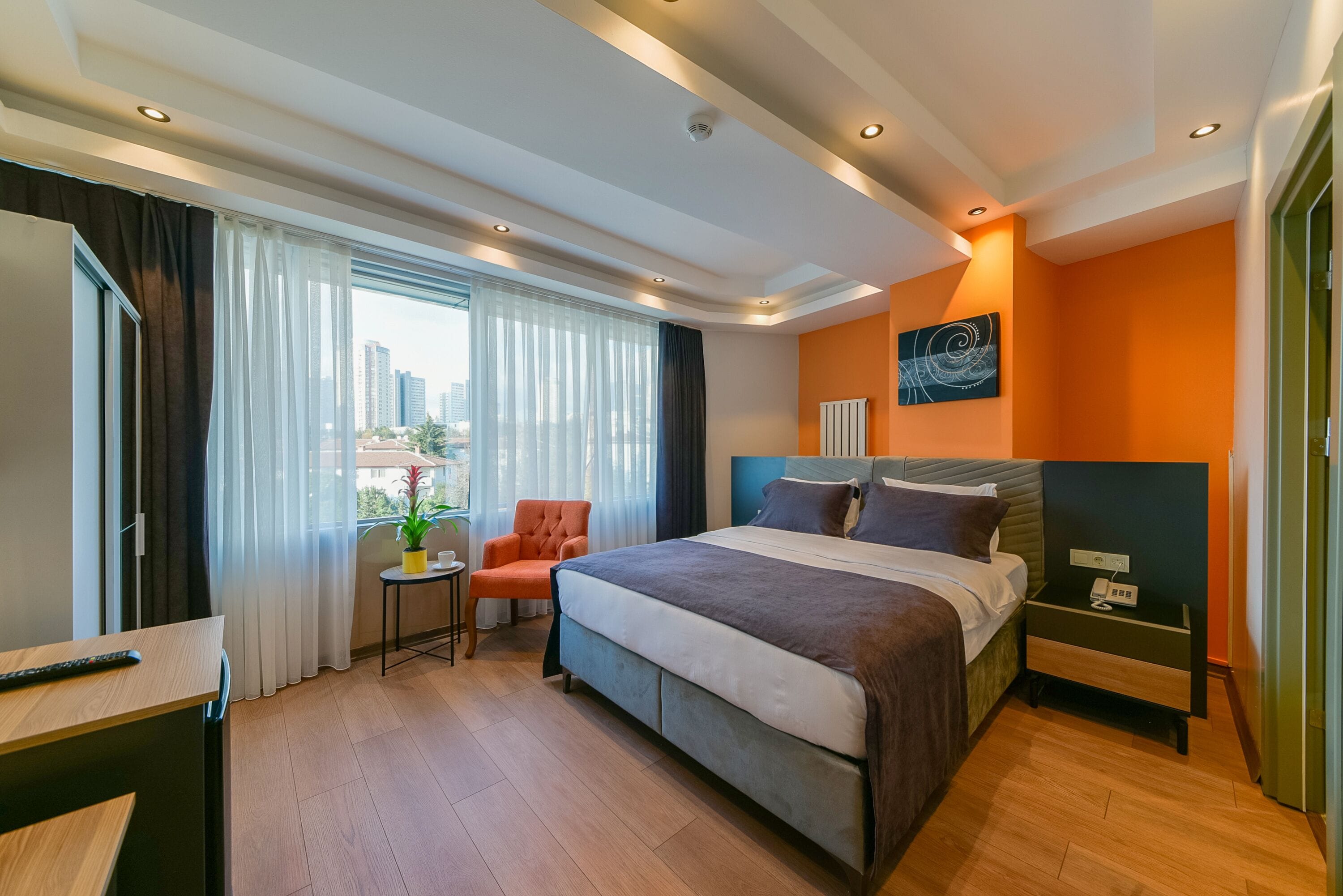 Una habitación de hotel moderna con una cama grande, una mesita con una silla, vistas a la ciudad a través de amplios ventanales y una mesita de noche con un teléfono.