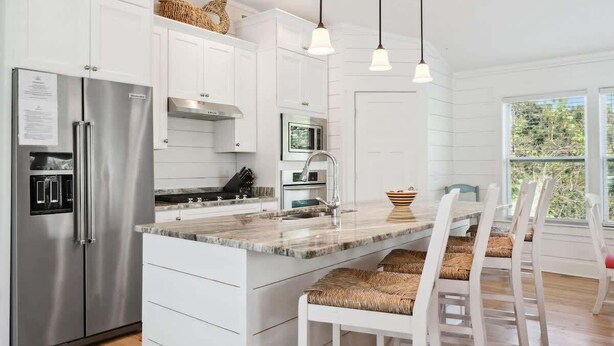 A modern kitchen with white cabinetry, stainless steel appliances, a marble countertop, and a dining area with white chairs.