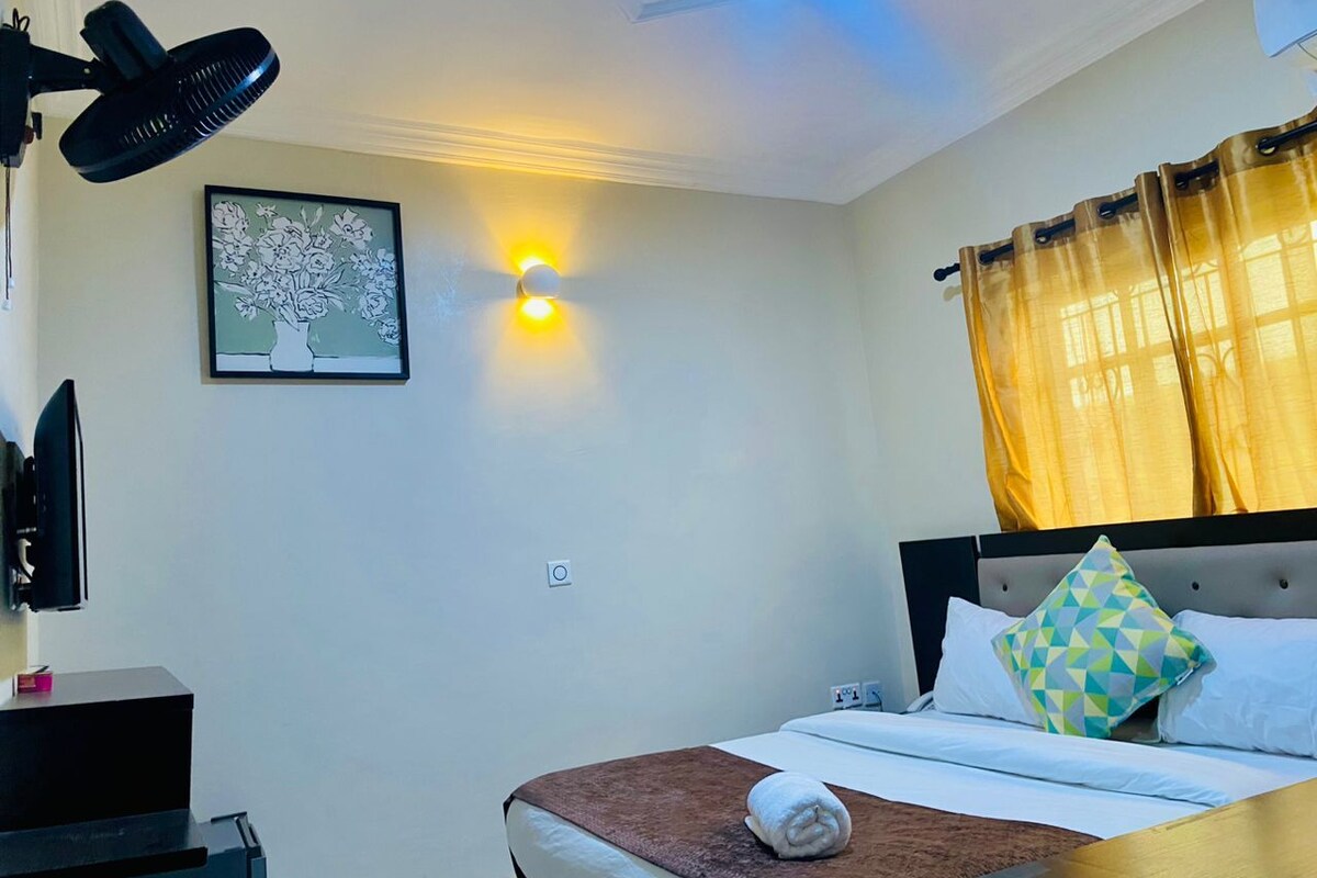 A hotel room with a bed, a bedside table, a TV, a wall-mounted light, and a window with curtains.