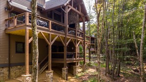 A two-story wooden cabin with a stone foundation, surrounded by a forest.