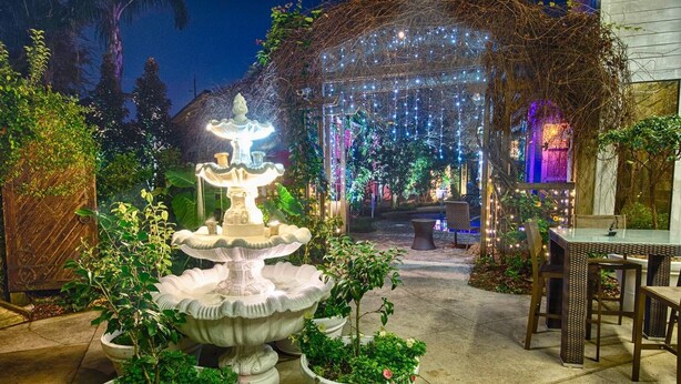 A well-lit outdoor patio area with a central fountain, surrounded by potted plants and a dining table with chairs.