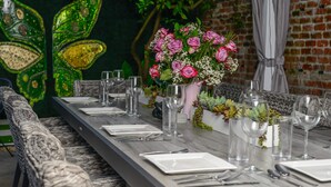 A long dining table set with plates, cutlery, and wine glasses, flanked by wicker chairs. A vase with pink flowers and greenery is placed on the table.