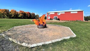 A circular fire pit with flames in the center, surrounded by a gravel area, with a red building in the background. - The Barndominium - Lots of Space! (Butternut)