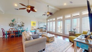 A spacious living room with a wooden floor, a large window with blinds, a ceiling fan, and a television. - Absolute Paradise: 3 BR, 2 BA House in Crystal Beach , Sleeps 14 (Bolivar Peninsula)