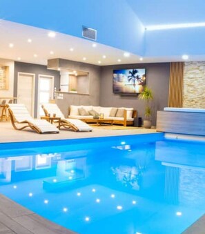 Ein moderner, überdachter Poolbereich mit Liegestühlen und einer Couch, umgeben von Wänden mit Steinverkleidung und indirekter Beleuchtung.