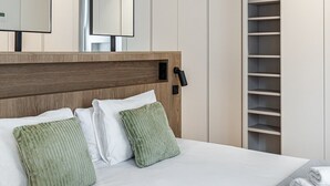 A modern hotel room with a large bed, two pillows, a bedside table, a mirror, and a built-in wardrobe.
