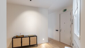 A hallway with a wooden floor, a black and wicker console table with a book and a glass, and a door with an emergency exit sign.