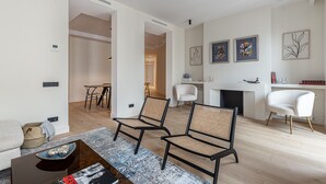 A modern living room with a sofa, two armchairs, a coffee table, and a fireplace. There are framed artworks on the wall and a small table with decorative items.