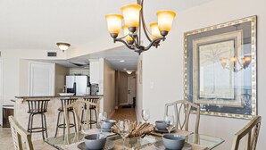 A dining area with a glass table, chairs, and a chandelier. There is a kitchen in the background. - Gulf-Front at Aquavista w/ Pool, Hot Tub & Sunset Views (Panama City Beach)