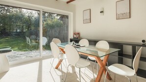 A dining area with a glass table and white chairs, wooden beams on the ceiling, and a view of the backyard through large sliding doors. - Idyllic Home in Picturesque Village of Wraysbury - Pass the Keys (Wraysbury)