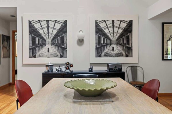 A dining area with a long table, two chairs, a decorative bowl, and two framed photographs on the wall.
