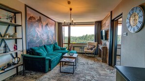Een moderne woonkamer met een groene bank, een salontafel en een groot schilderij van bergen. Er hangt een klok aan de muur en een televisie is aan de wand bevestigd.