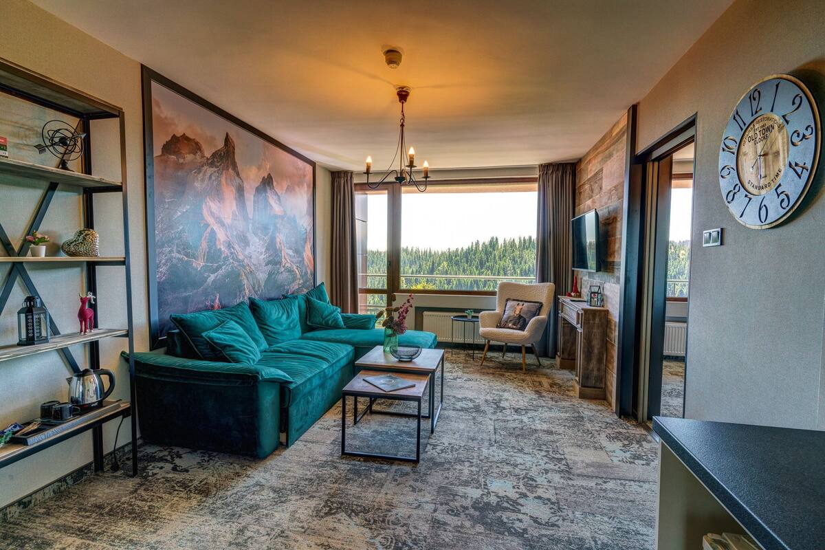 A modern living room with a green sofa, a coffee table, and a large painting of mountains. There is a clock on the wall and a TV mounted on the wall.