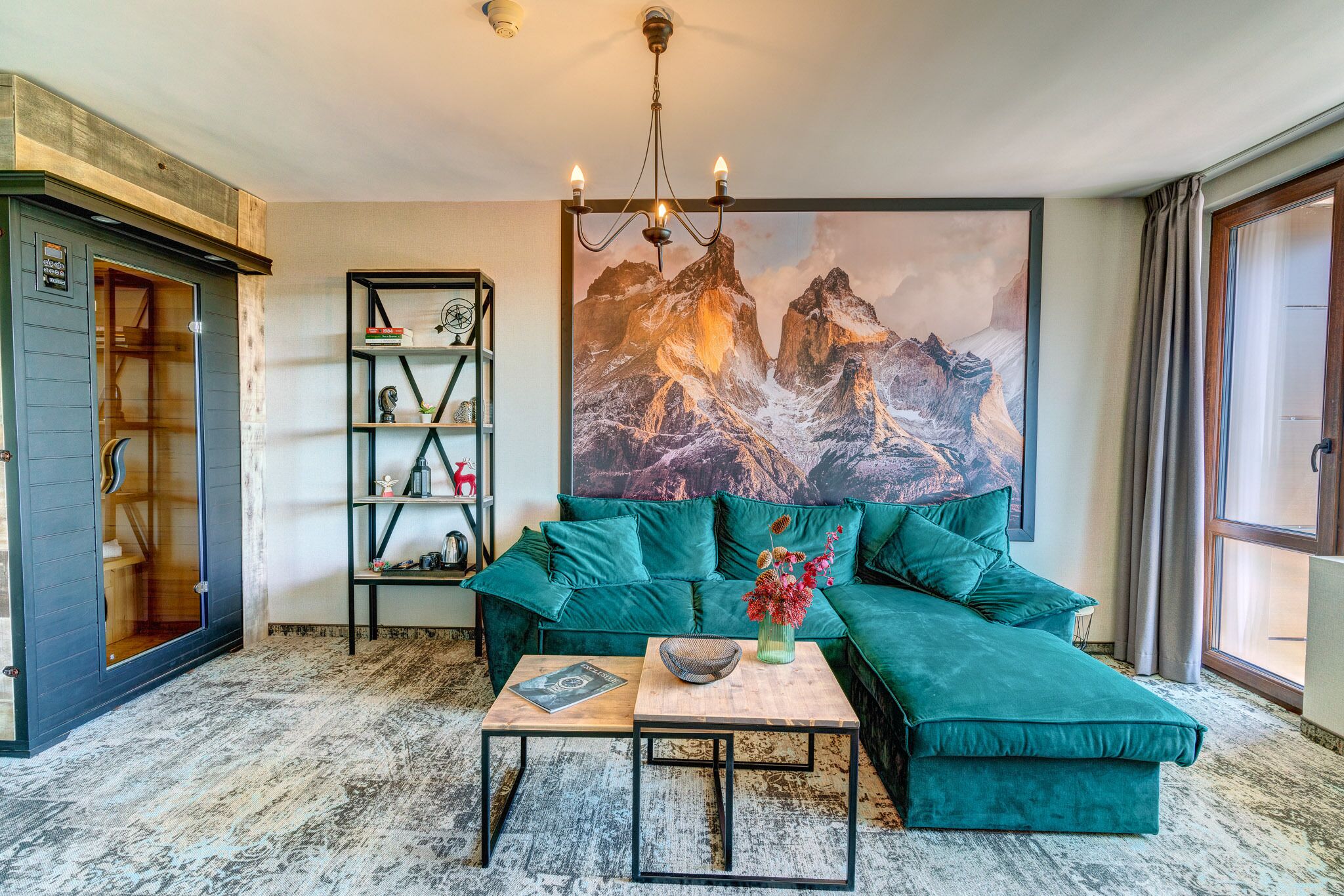 A living room with a green sectional sofa, a coffee table, a bookshelf, and a large mountain painting.