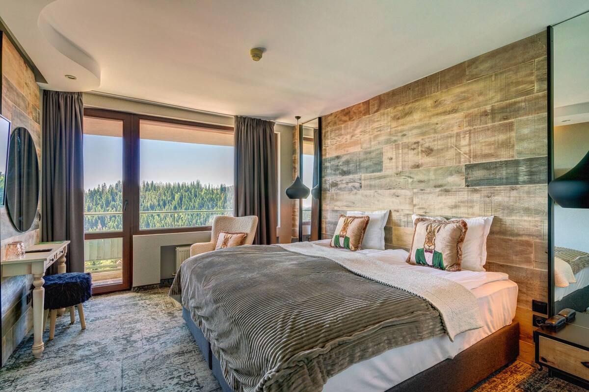 A modern hotel room with a large bed, a desk, and a view of the forest.