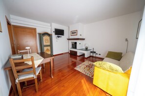 Una sala de estar con piso de madera, una mesa de comedor con sillas, un sofá, un televisor montado en la pared y una chimenea.