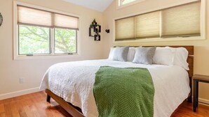Een slaapkamer met een groot bed, twee ramen met jaloezieën, een nachtkastje en een groene plaid.