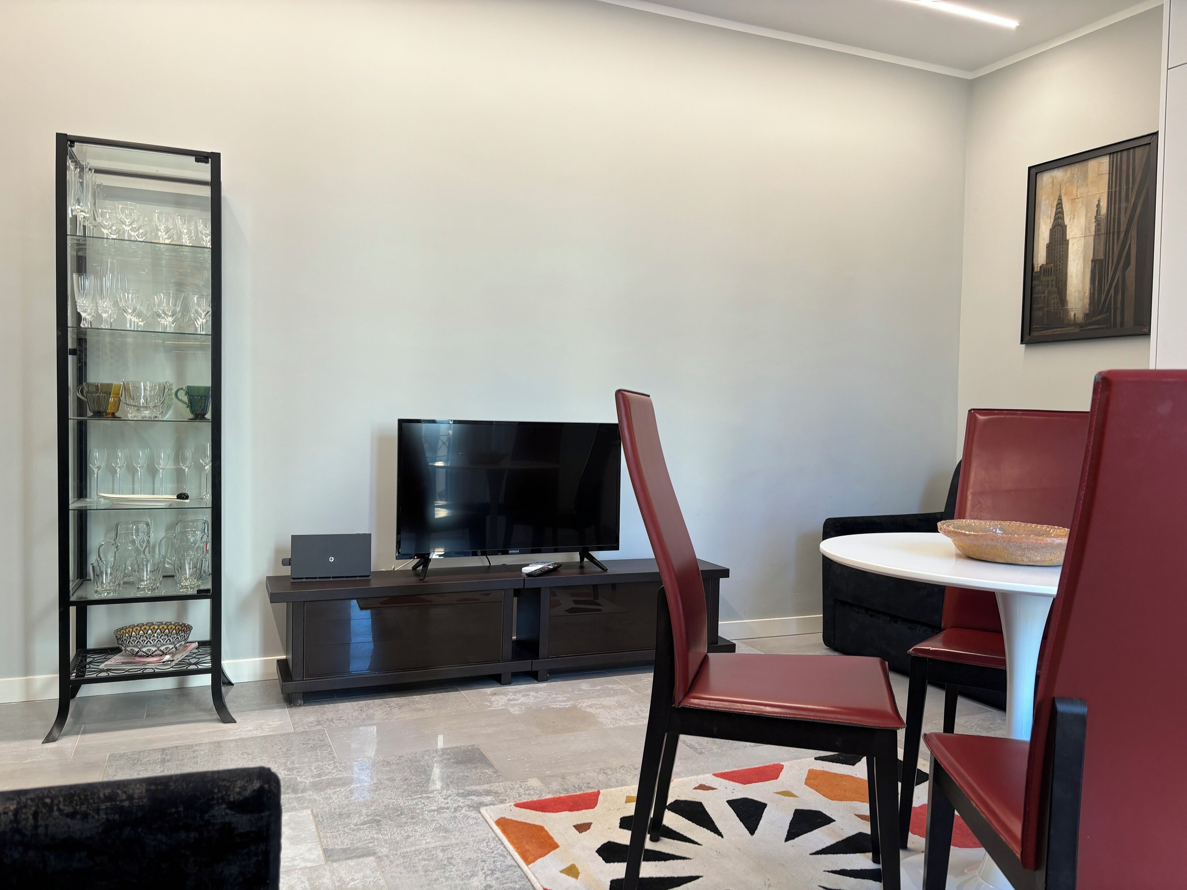 A modern living room with a flat-screen TV, a glass display cabinet, and a dining area with red chairs and a white table.
