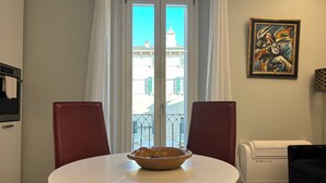 Ein Essbereich mit einem runden Tisch, zwei roten Stühlen, einer Schale auf dem Tisch, einem Fenster mit weißen Vorhängen und einem Gemälde an der Wand.