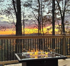 A deck with a glass-top fire pit surrounded by a wooden railing, overlooking a forested area during sunset. - Tiny home BIG View!  (Knoxville)