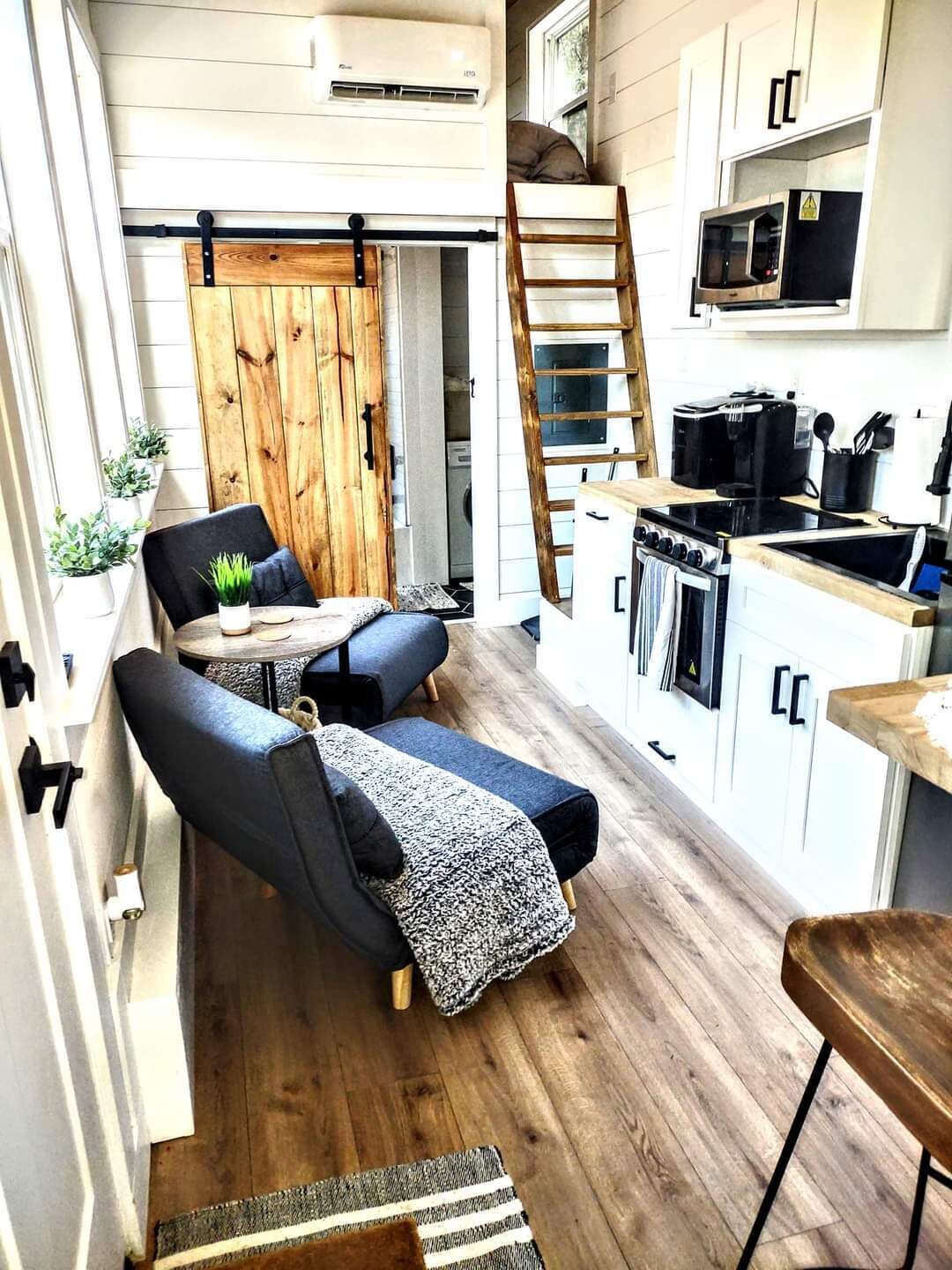 A compact kitchen with a stove, microwave, and sink. There is a wooden ladder leading to a lofted sleeping area.