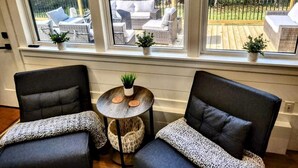 Une pièce comprenant deux fauteuils sombres, une petite table ronde ornée d’une plante, et donnant sur une terrasse extérieure meublée de mobilier en osier.