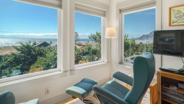 A room with a large window overlooking a beach, a television, and a bookshelf.