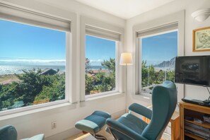 A room with a large window overlooking a beach, a television, and a bookshelf.