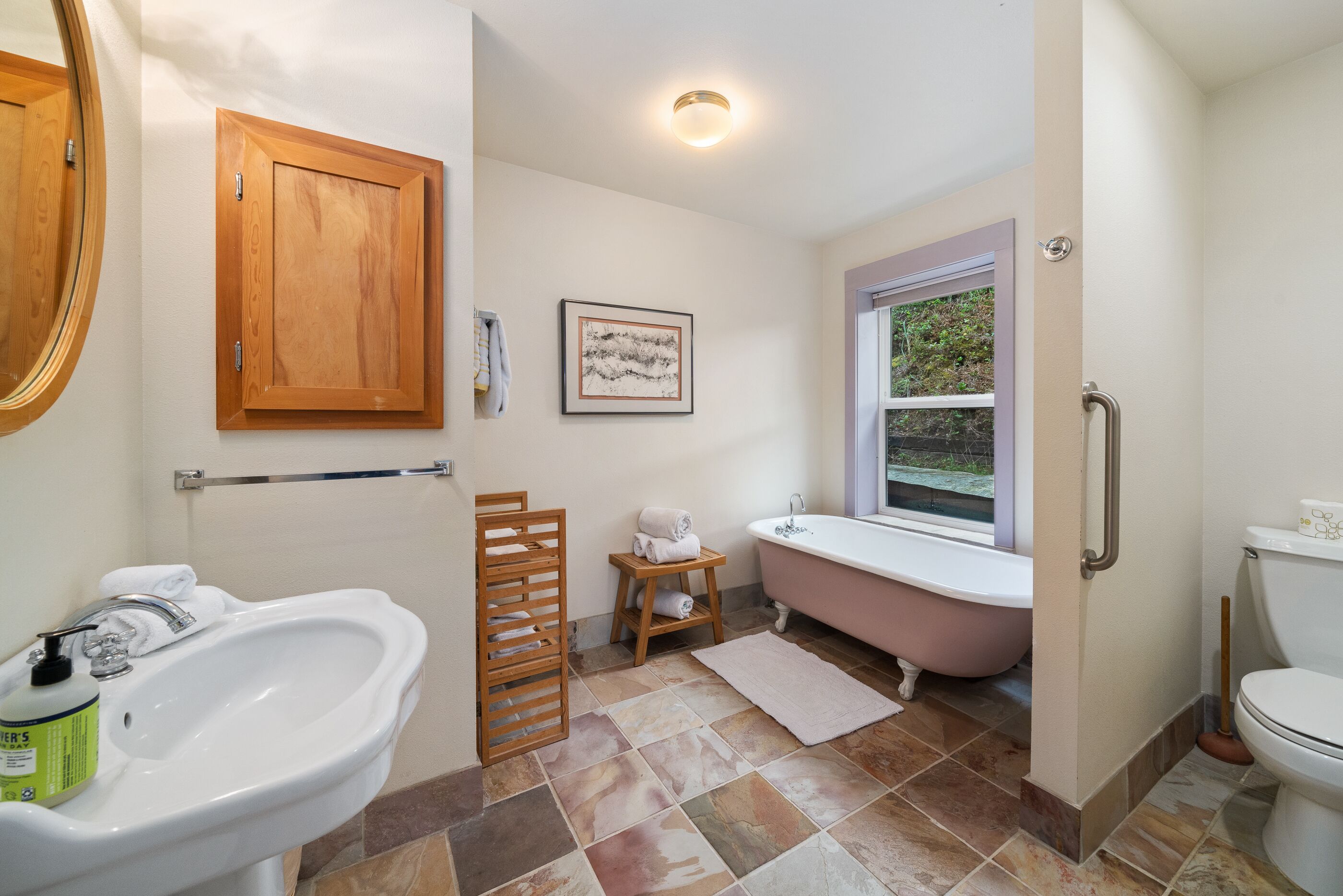 Un baño con lavamanos blanco, mueble de madera, una obra de arte enmarcada, una bañera rosa y un inodoro.