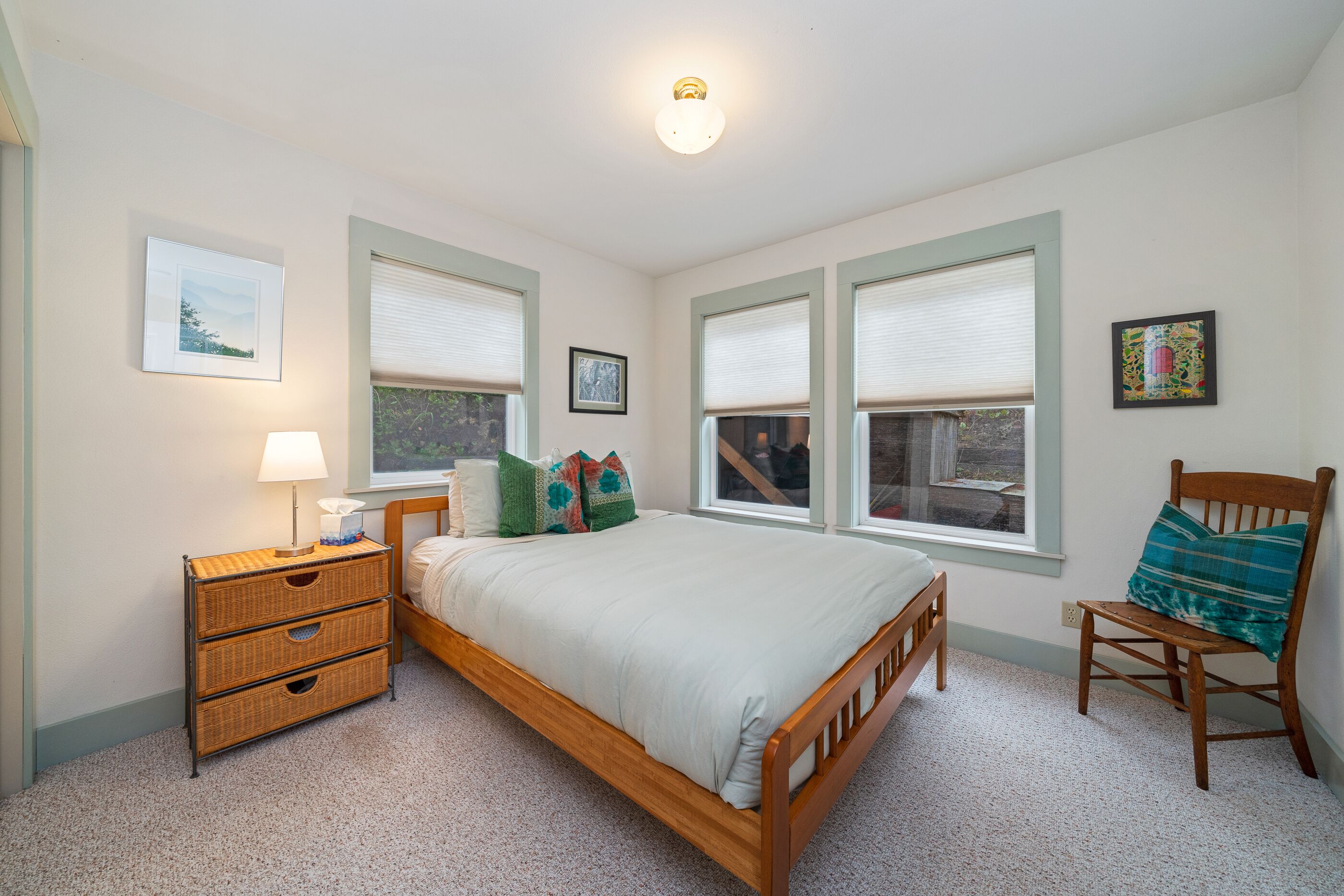 A bedroom with a single bed, a nightstand with a lamp, a chair, and two windows with blinds.