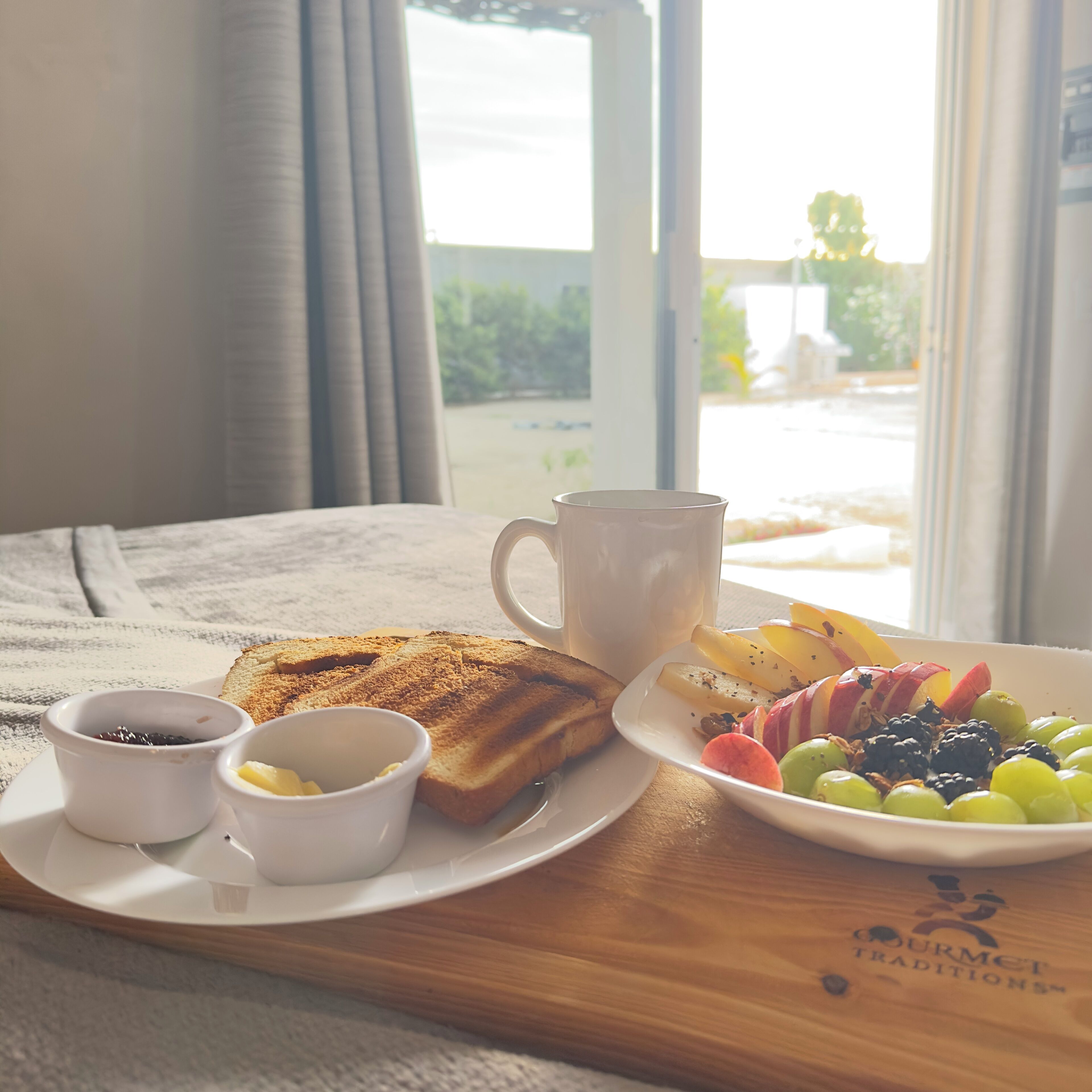 Een ontbijtplateau met toast, een mok en een bord met fruit en bessen op een houten tafel bij een raam.