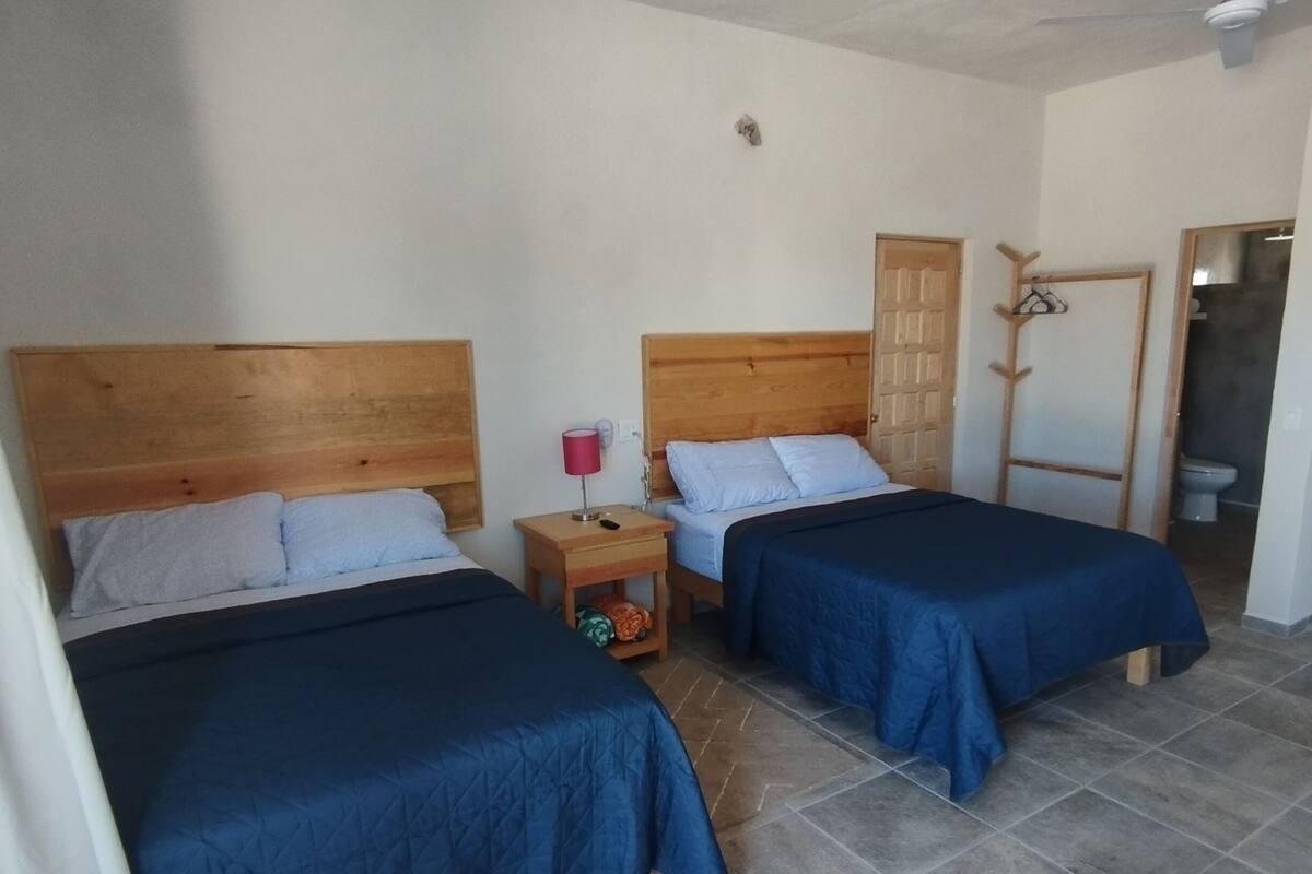 Ein Zimmer mit zwei Betten, einem Holz-Kopfteil, einem Nachttisch, einer roten Lampe und einer Tür mit Kleiderhaken.