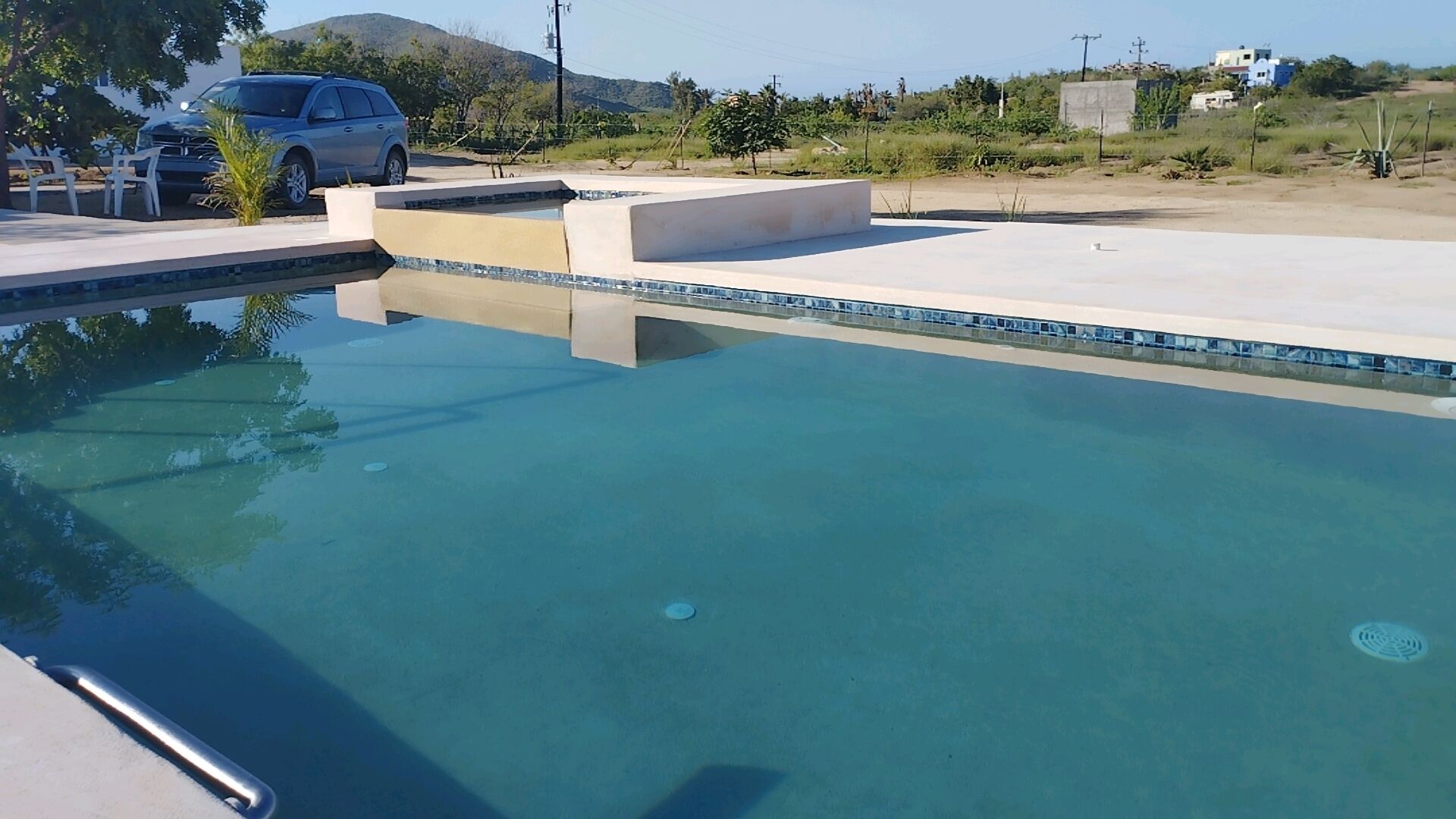 Une piscine à l’eau d’un bleu limpide, avec une terrasse en béton et une voiture garée à proximité.