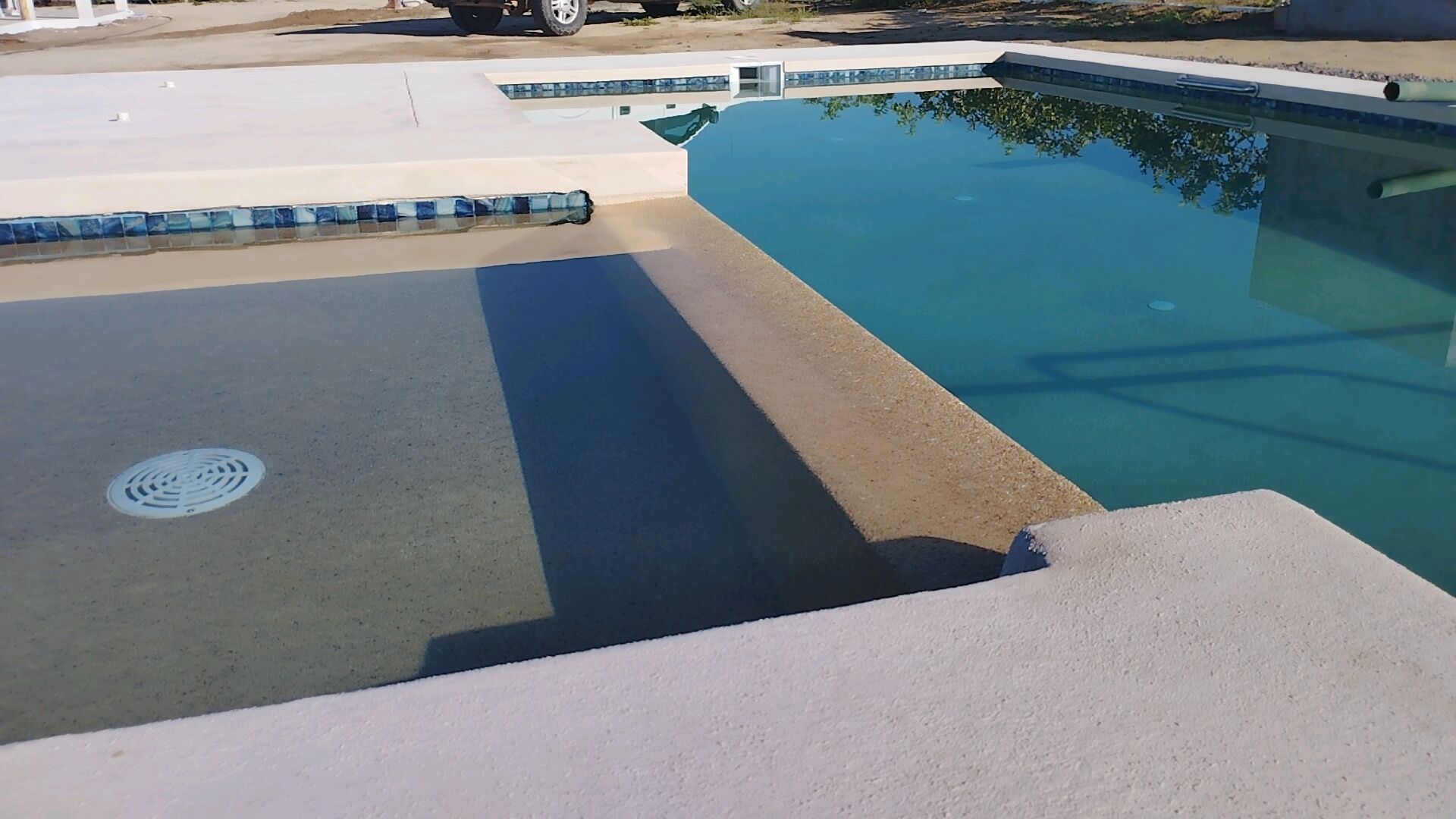 A swimming pool with a clear blue water surface, a concrete deck, and a manhole cover.
