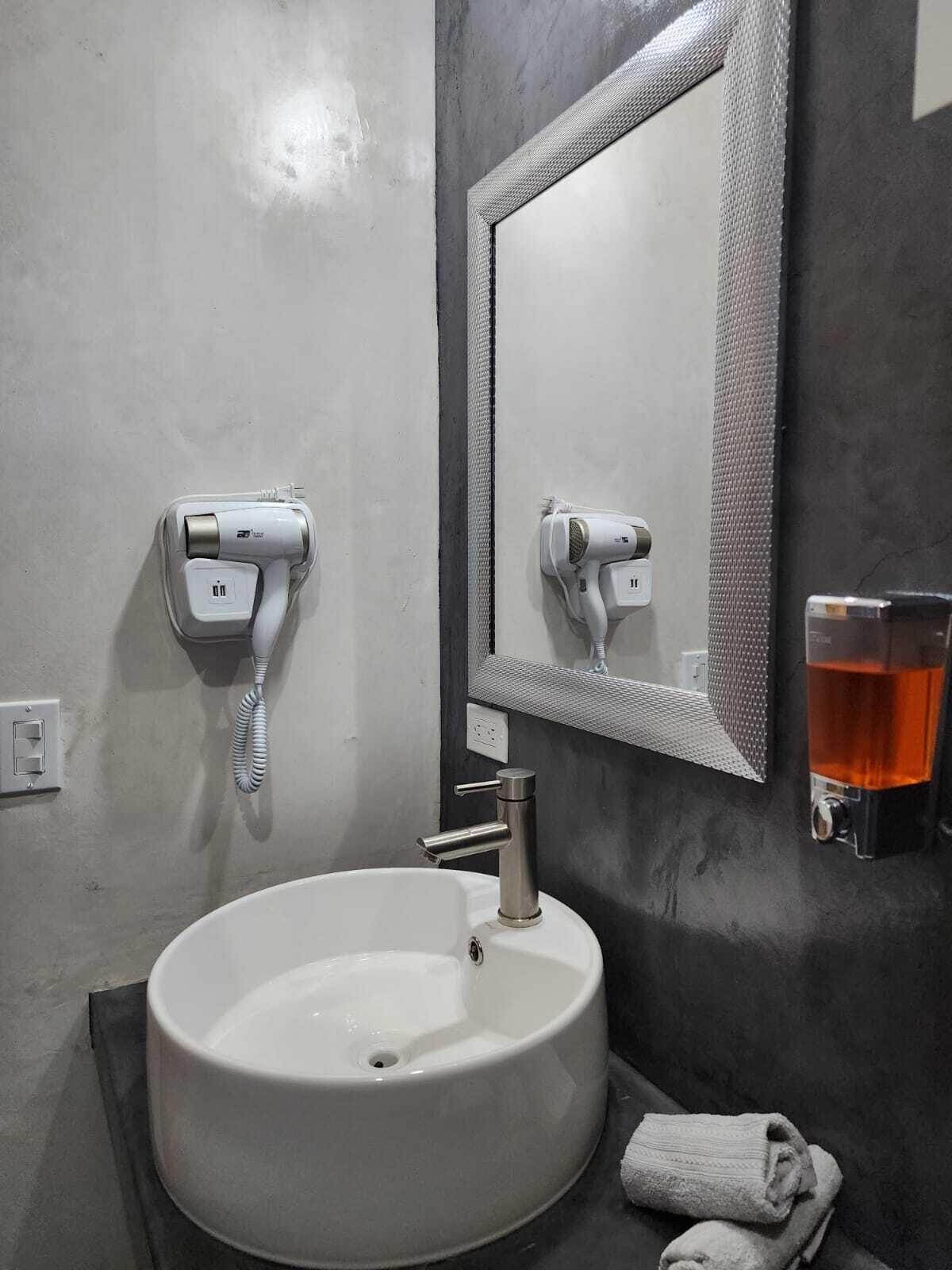 Un baño con un lavamanos blanco, un espejo, un secador de pelo y un vaso de agua.