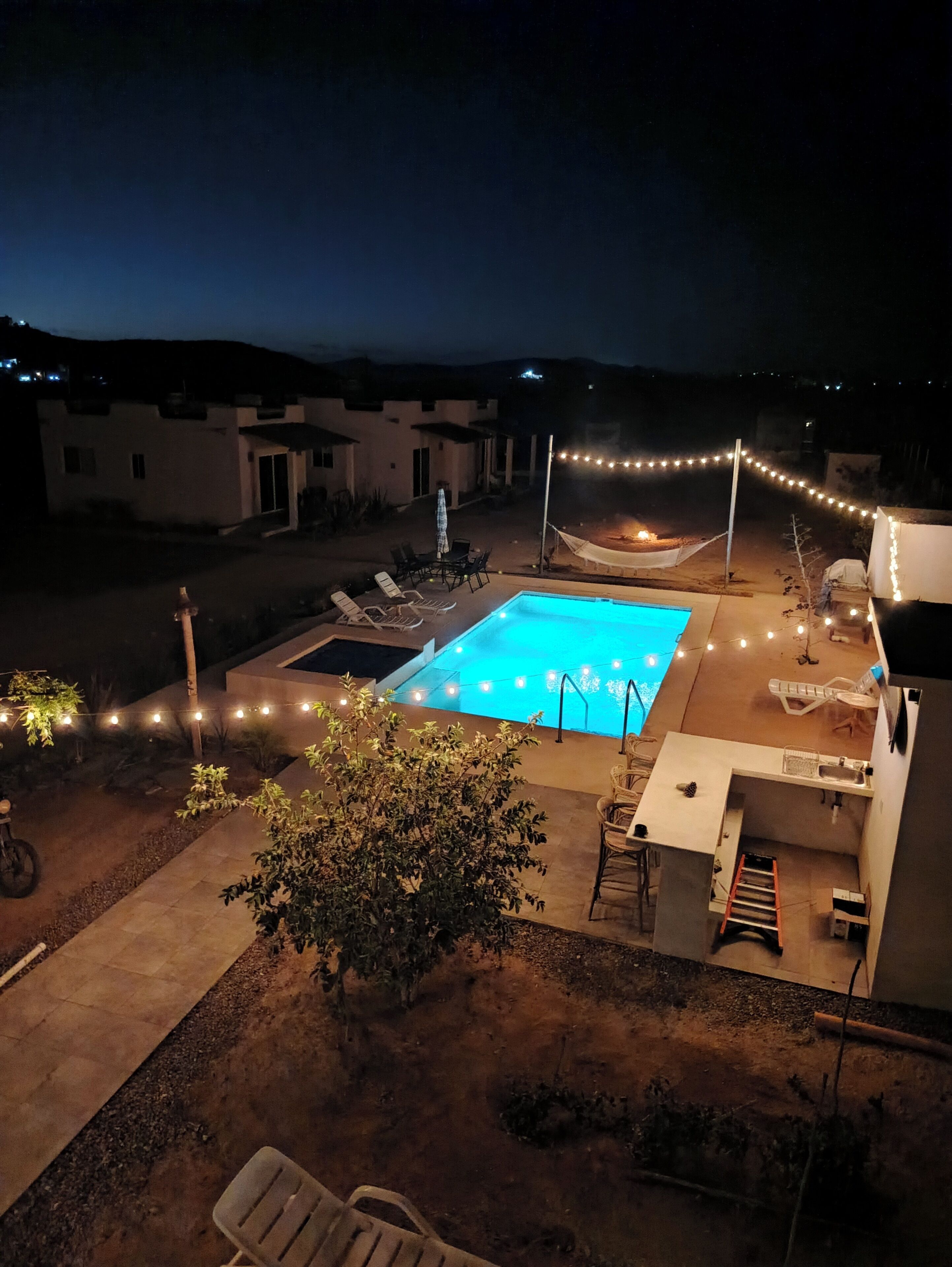 A swimming pool surrounded by outdoor seating and string lights, with a view of a hillside in the background.