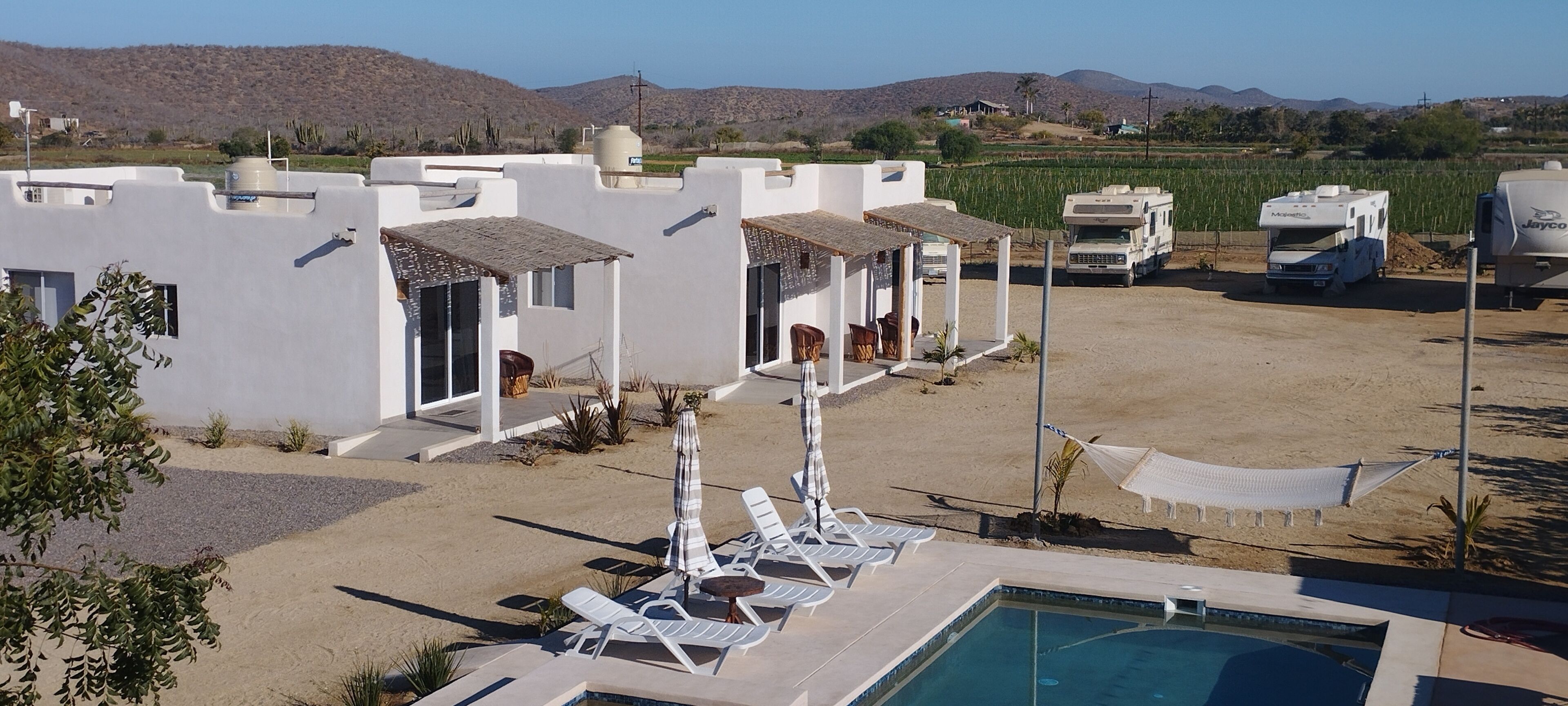 Un complejo turístico con edificios blancos, una piscina y vehículos recreativos al fondo.