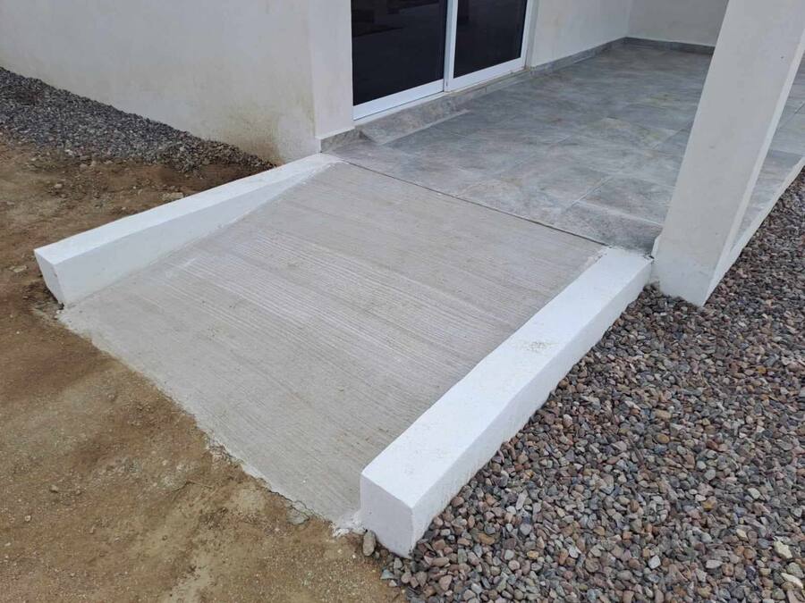 Eine Betonrampe führt zu einem Gebäudeeingang, daneben ein Kiesweg.