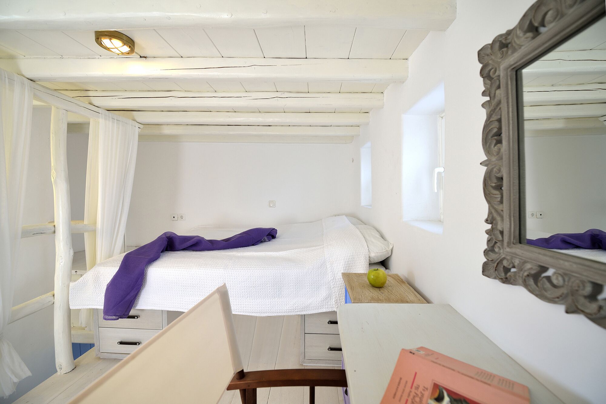 Una habitación pequeña y funcional con una cama, una silla de madera, una mesita con una manzana y un espejo en la pared.