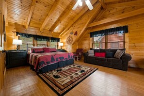 Interior de una cabaña de troncos con techo de madera, una cama con colcha roja y negra, un sofá marrón oscuro y una alfombra estampada.