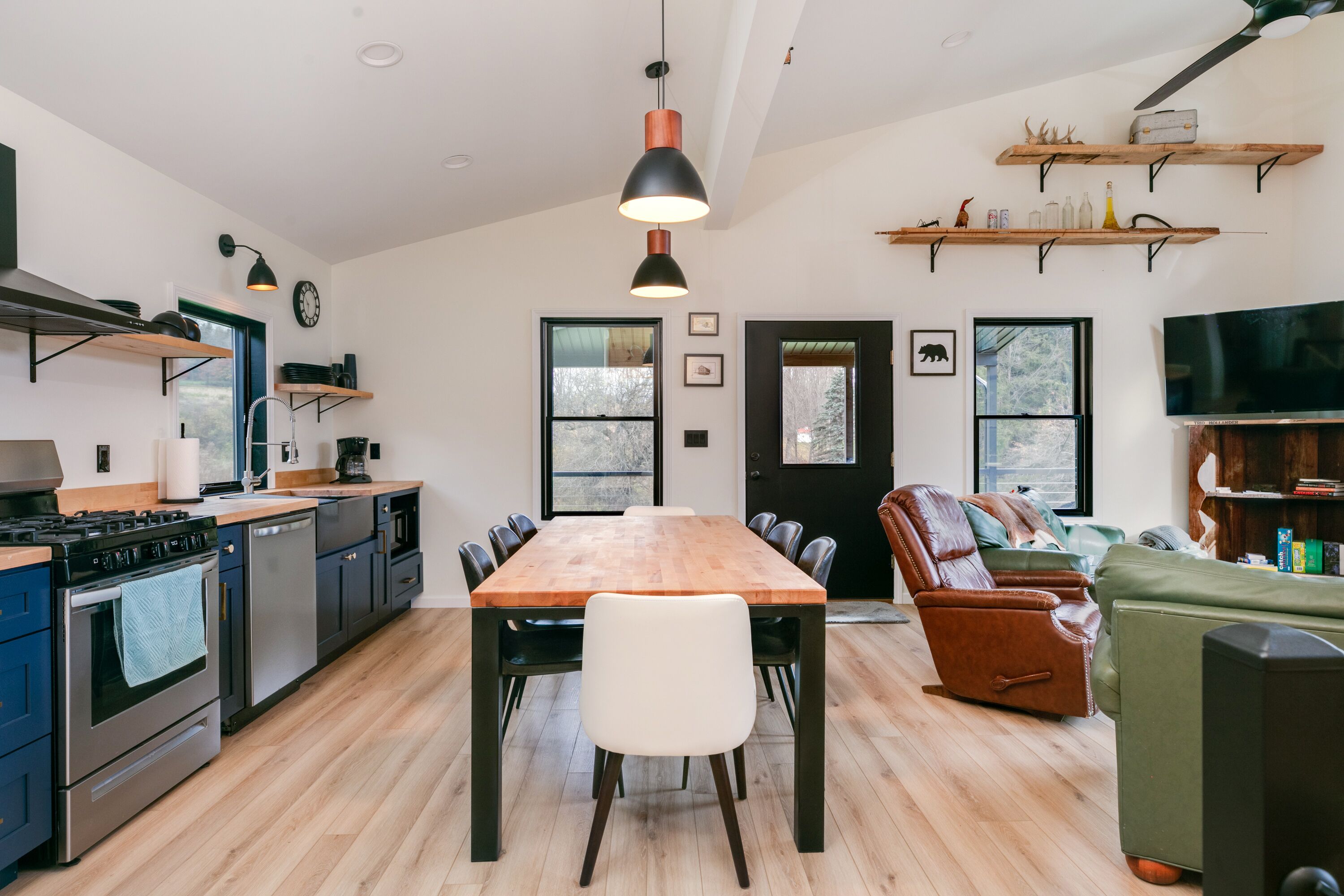 Une cuisine moderne avec une table à manger en bois, des chaises noires et un grand téléviseur. On trouve des étagères en bois au-dessus de la table et un canapé en cuir dans l’espace salon.