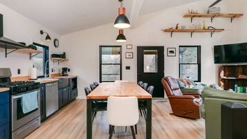Une cuisine moderne avec une table à manger en bois, des chaises noires et un grand téléviseur. On trouve des étagères en bois au-dessus de la table et un canapé en cuir dans l’espace salon.