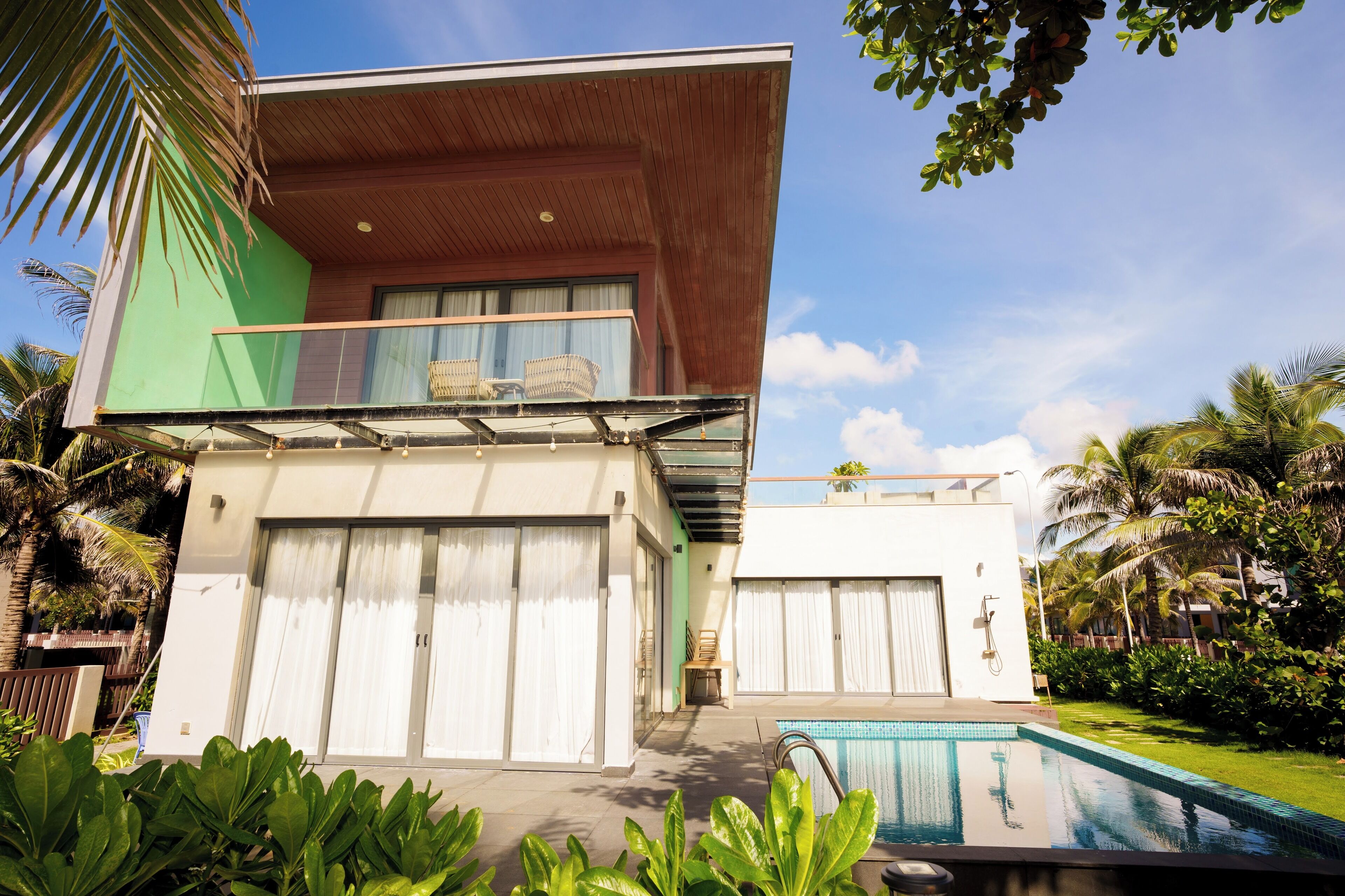 A modern two-story house with a balcony, a swimming pool, and lush greenery.