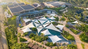 Vue aérienne d’un complexe hôtelier doté d’une piscine centrale, entourée de palmiers et surmontée de toiles blanches.