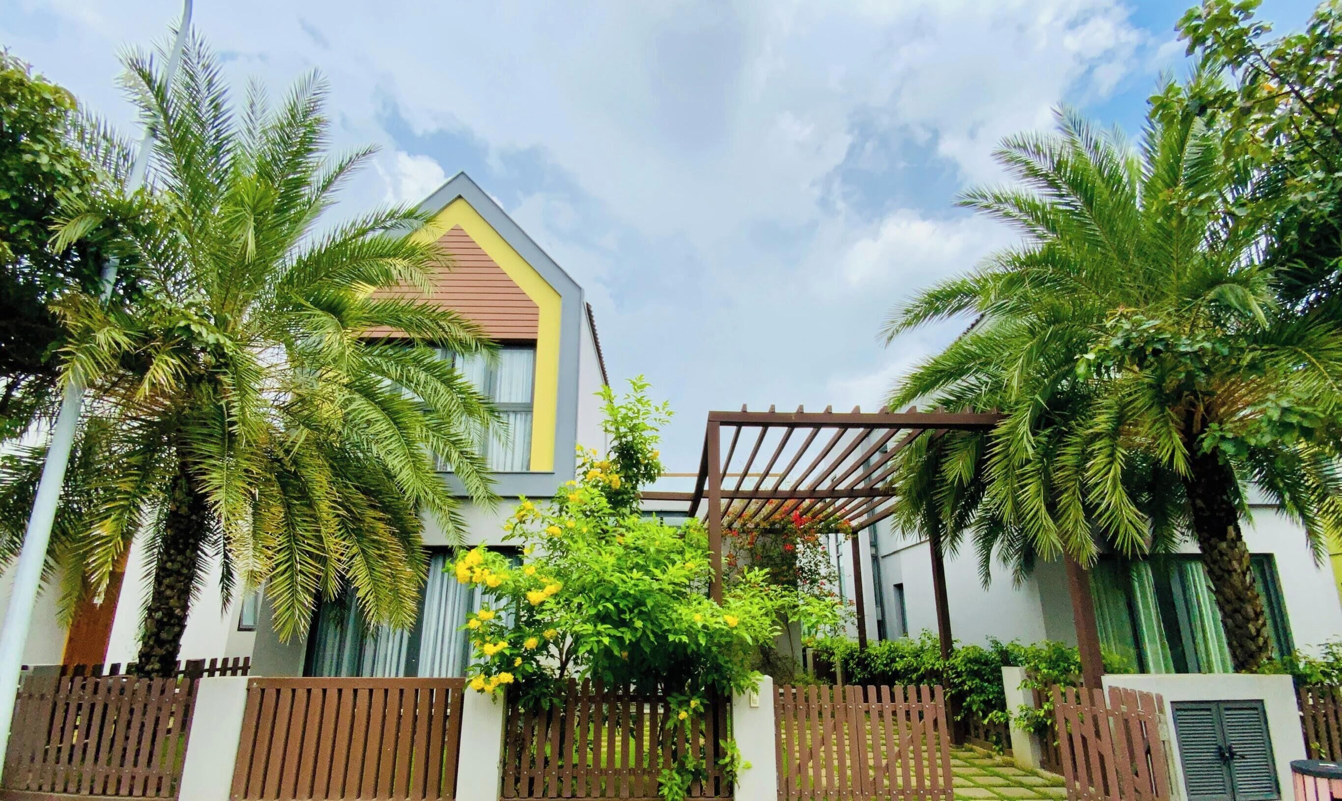 A modern two-story house with a sloped roof, surrounded by lush greenery and palm trees.
