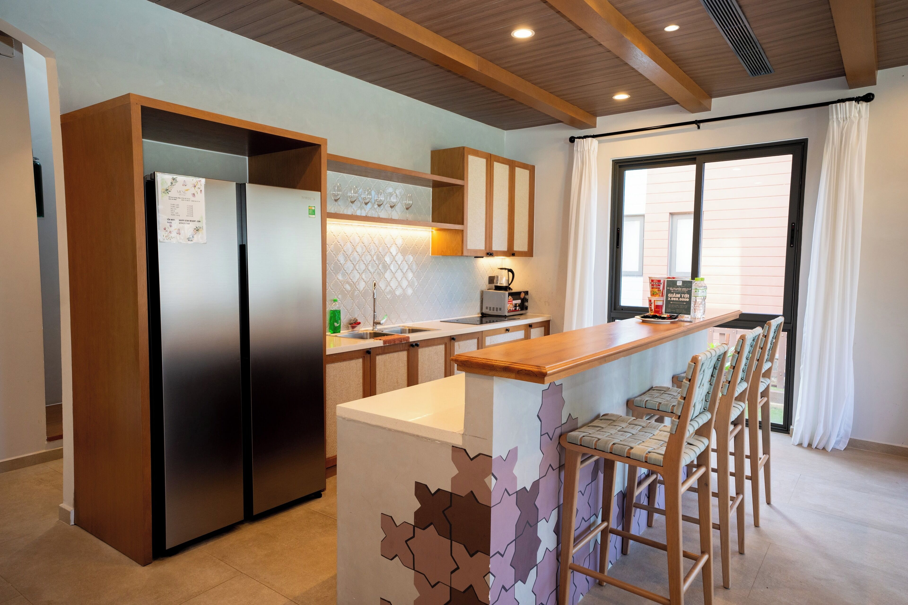 A modern kitchen with a built-in refrigerator, wooden cabinets, a bar area with bar stools, and a large window with curtains.