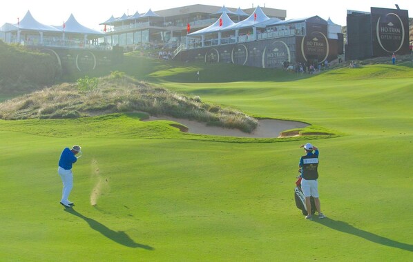 Un campo de golf con un putting green, búnkeres de arena y un club de campo al fondo.