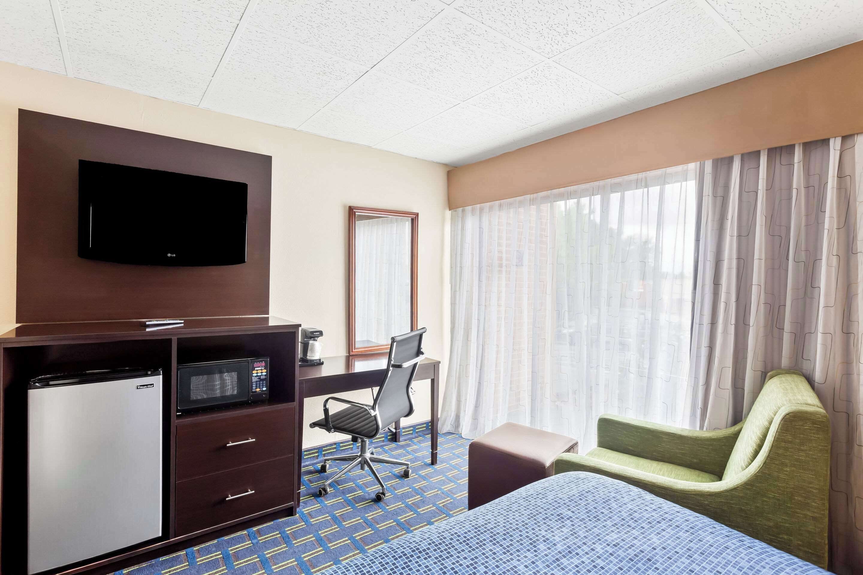 Habitación de hotel con cama, escritorio con computadora, refrigerador, microondas, televisión y ventana con cortinas.