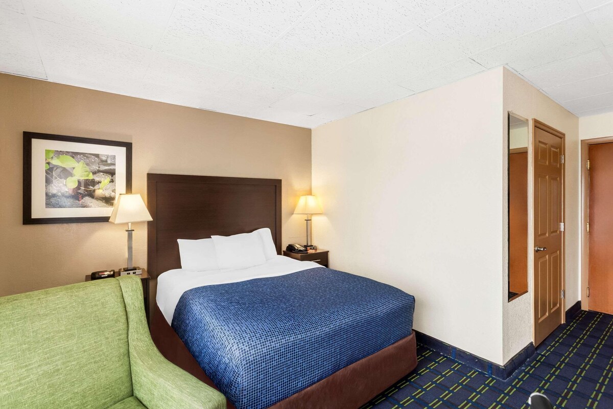 Habitación de hotel con cama, dos lámparas de noche, un sillón verde, un cuadro en la pared y una puerta que da a otra habitación.