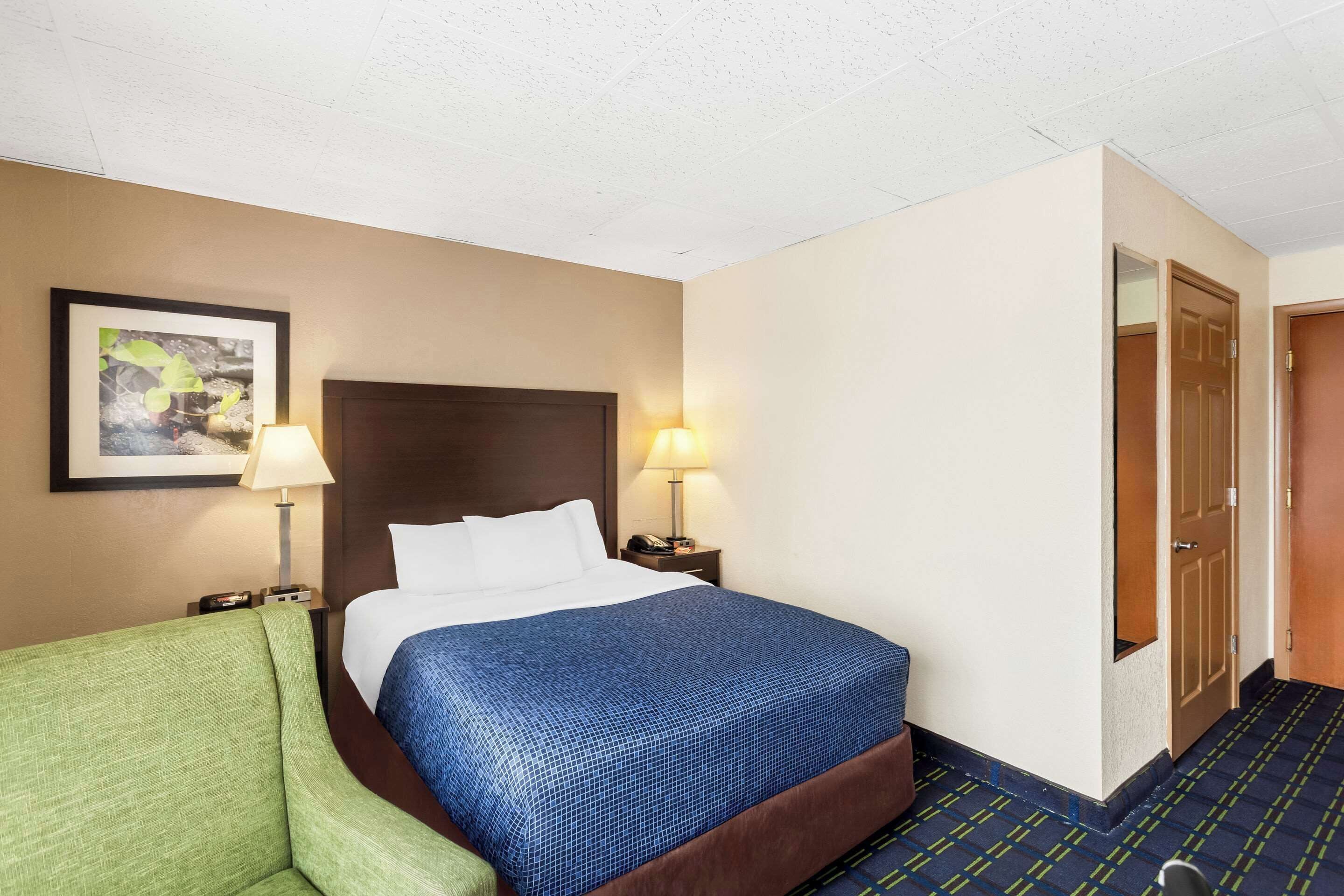 A hotel room with a bed, two bedside lamps, a green armchair, a framed picture on the wall, and a door to another room.